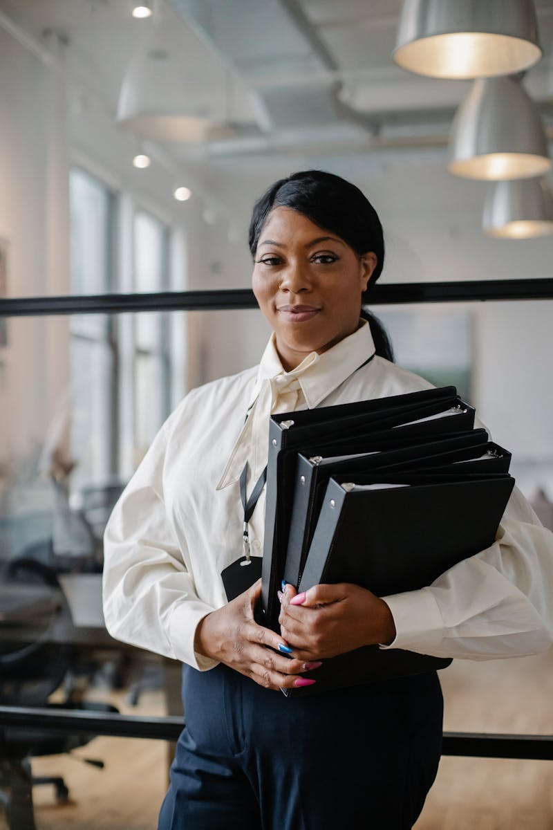 Photo by Sora Shimazaki Smiling African American female employee wearing classy outfit standing with paper documents in modern workplace